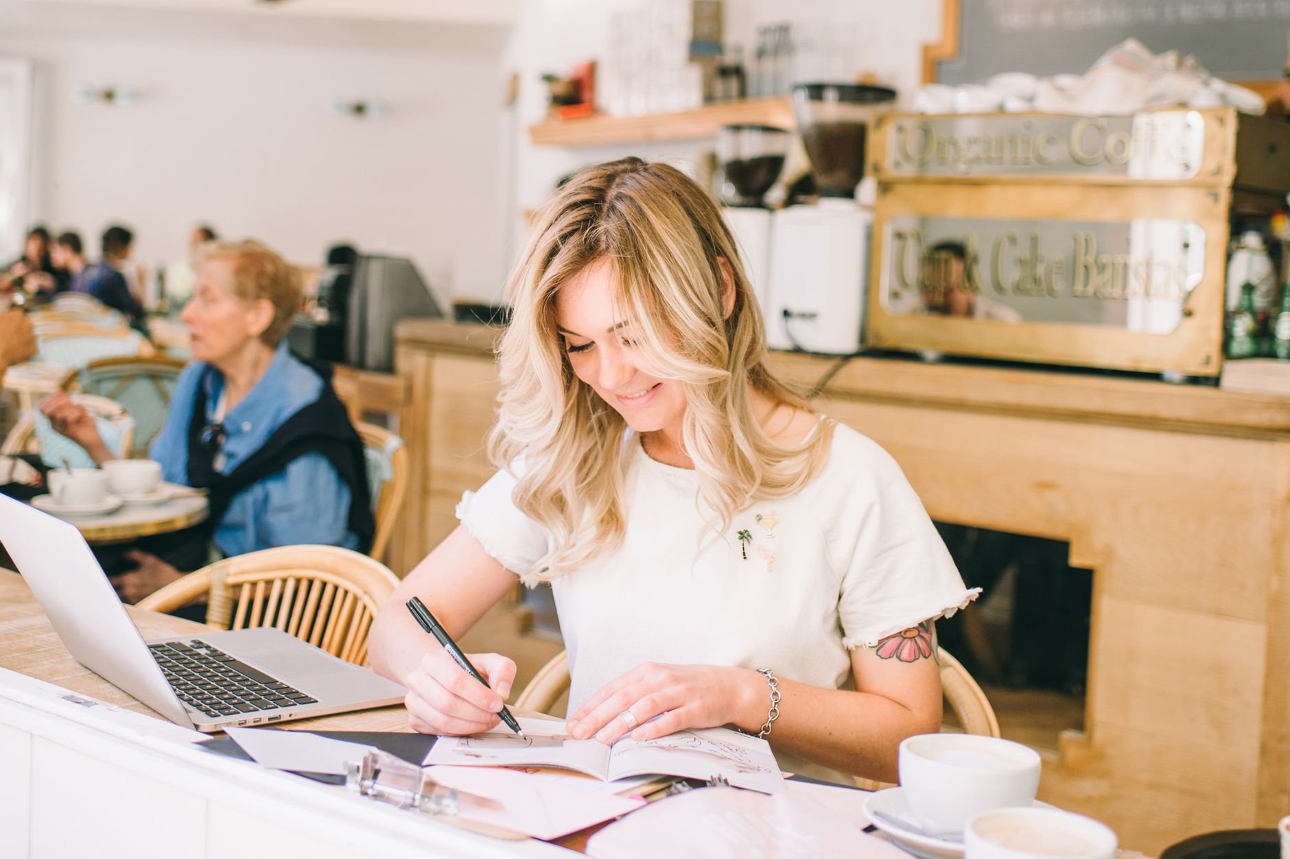 A woman working on her laptop and writing in a bright café, embodying a relaxed freelance lifestyle.